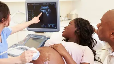 Ultrasound scan in pregnancy, woman lying on hospital bed with partner, doctor pointing at monitor.